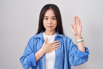 Young chinese woman standing over white background swearing with hand on chest and open palm, making a loyalty promise oath