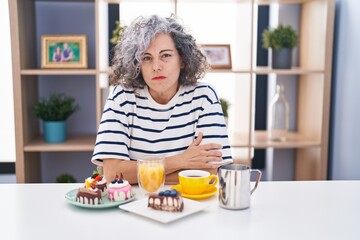 Middle age woman with grey hair eating pastries and drinking coffee for breakfast skeptic and nervous, disapproving expression on face with crossed arms. negative person.