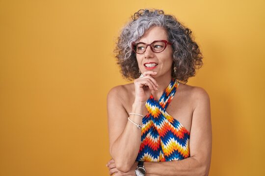 Middle age woman with grey hair standing over yellow background with hand on chin thinking about question, pensive expression. smiling and thoughtful face. doubt concept.