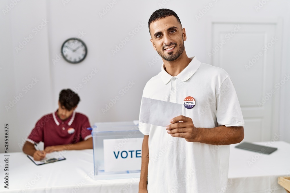 Poster Young hispanic man voting putting envelop in ballot box looking positive and happy standing and smiling with a confident smile showing teeth