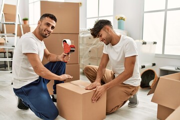 Two hispanic men couple smiling confident packing cardboard box at new home