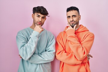 Young hispanic gay couple standing over pink background looking confident at the camera smiling with crossed arms and hand raised on chin. thinking positive.