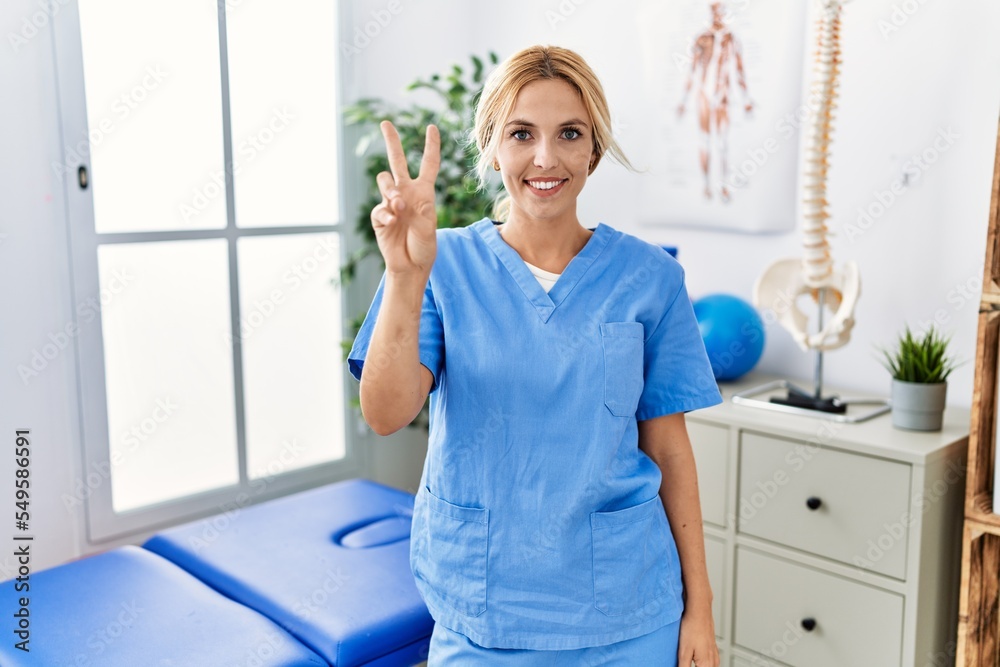 Poster Beautiful blonde physiotherapist woman working at pain recovery clinic showing and pointing up with fingers number two while smiling confident and happy.