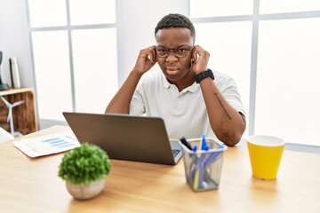 Young african man working at the office using computer laptop covering ears with fingers with...