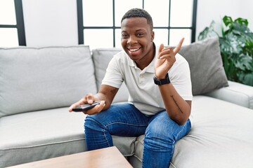 Young african man watching tv holding television remote control smiling happy pointing with hand and finger to the side