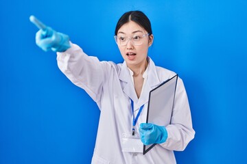 Chinese young woman working at scientist laboratory pointing with finger surprised ahead, open mouth amazed expression, something on the front