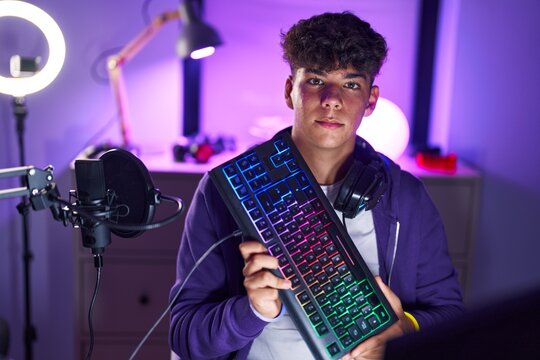 Young hispanic teenager streamer holding computer keyboard with relaxed expression at gaming room