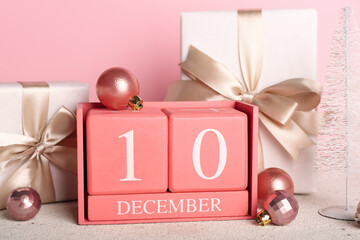 Cube calendar with date 10 DECEMBER, Christmas balls and gifts on table near pink wall