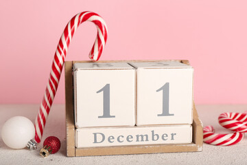Cube calendar with date 11 DECEMBER, candy canes and Christmas balls on table near pink wall