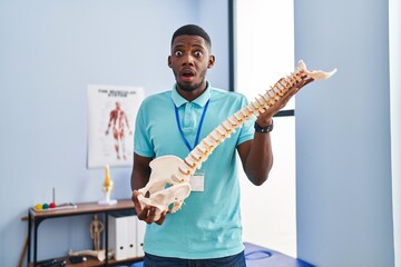African american man holding anatomical model of spinal column afraid and shocked with surprise and amazed expression, fear and excited face.