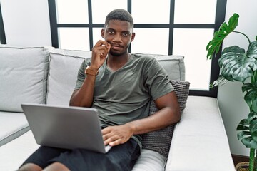 Young african american man using laptop at home sitting on the sofa mouth and lips shut as zip with...