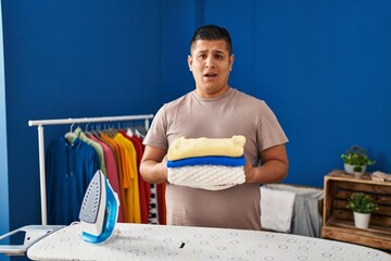 Hispanic young man holding folded laundry after ironing clueless and confused expression. doubt...