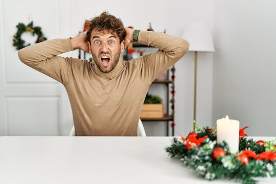 Young handsome man with beard sitting on the table by christmas decoration crazy and scared with hands on head, afraid and surprised of shock with open mouth