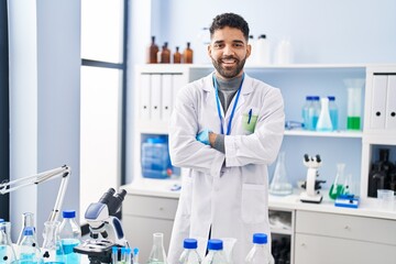 Young hispanic man wearing scientist uniform standing with arms crossed gesture at laboratory