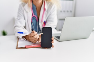 Middle age blonde woman wearing doctor uniform showing smartphone screen at clinic