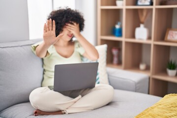 Young brunette woman with curly hair using laptop sitting on the sofa at home covering eyes with hands and doing stop gesture with sad and fear expression. embarrassed and negative concept.