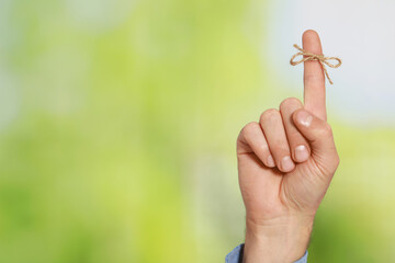 Man showing index finger with tied bow as reminder on green blurred background, closeup. Space for...