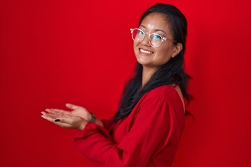 Asian young woman standing over red background pointing aside with hands open palms showing copy space, presenting advertisement smiling excited happy