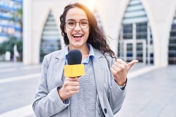 Young brunette woman holding reporter microphone pointing thumb up to the side smiling happy with open mouth