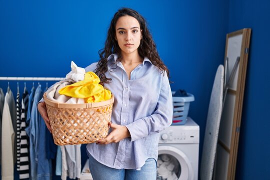 Young brunette woman holding laundry basket thinking attitude and sober expression looking self confident