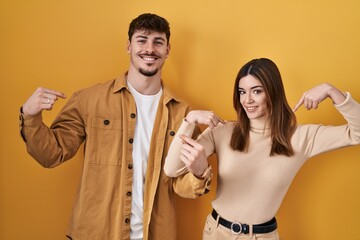 Young hispanic couple standing over yellow background looking confident with smile on face, pointing oneself with fingers proud and happy.