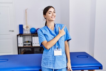 Young hispanic woman wearing physiotherapist uniform standing at clinic cheerful with a smile on face pointing with hand and finger up to the side with happy and natural expression
