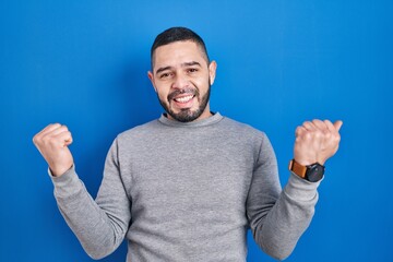 Fototapeta premium Hispanic man standing over blue background celebrating surprised and amazed for success with arms raised and eyes closed. winner concept.