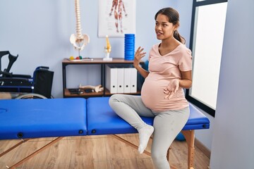 Young latin woman pregnant patient sitting on massage table at clinic