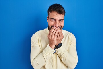 Handsome hispanic man standing over blue background laughing and embarrassed giggle covering mouth...