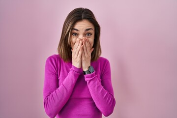 Hispanic woman standing over pink background laughing and embarrassed giggle covering mouth with...