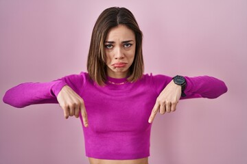 Hispanic woman standing over pink background pointing down looking sad and upset, indicating direction with fingers, unhappy and depressed.