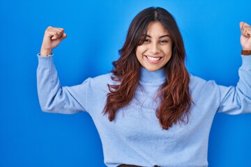 Hispanic young woman standing over blue background showing arms muscles smiling proud. fitness concept.