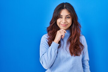 Hispanic young woman standing over blue background with hand on chin thinking about question, pensive expression. smiling and thoughtful face. doubt concept.