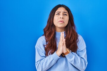 Hispanic young woman standing over blue background begging and praying with hands together with hope expression on face very emotional and worried. begging.