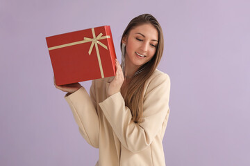 Young woman in blazer with Christmas gift on lilac background