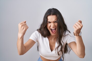Young teenager girl standing over white background angry and mad raising fists frustrated and furious while shouting with anger. rage and aggressive concept. - obrazy, fototapety, plakaty