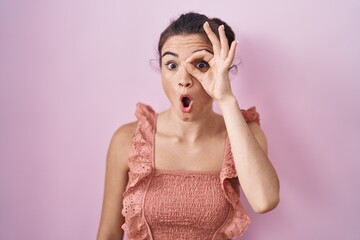 Young teenager girl standing over pink background doing ok gesture shocked with surprised face, eye looking through fingers. unbelieving expression.