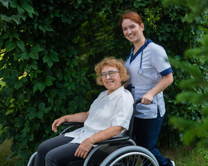 Caucasian female doctor walks with an elderly patient in a wheelchair in the park. Nurse accompanies an old woman on a walk outdoors. 