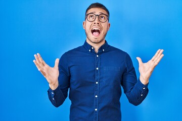 Fototapeta premium Young hispanic man wearing glasses over blue background crazy and mad shouting and yelling with aggressive expression and arms raised. frustration concept.