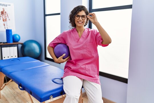 Young latin woman wearing physiotherapist uniform holding fit ball at clinic