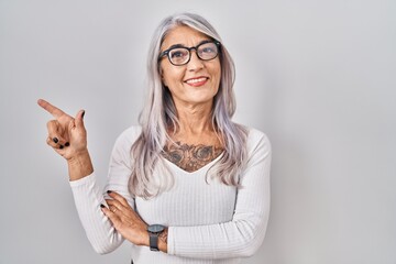 Middle age woman with grey hair standing over white background with a big smile on face, pointing with hand finger to the side looking at the camera.