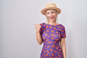 Young caucasian woman wearing flowers dress and summer hat smiling with happy face looking and pointing to the side with thumb up.