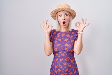 Young caucasian woman wearing flowers dress and summer hat looking surprised and shocked doing ok approval symbol with fingers. crazy expression