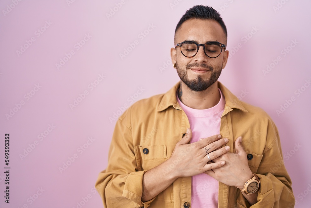 Poster Young hispanic man standing over pink background smiling with hands on chest with closed eyes and grateful gesture on face. health concept.