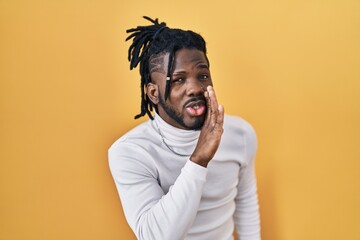African man with dreadlocks wearing turtleneck sweater over yellow background hand on mouth telling secret rumor, whispering malicious talk conversation - obrazy, fototapety, plakaty