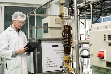 Apothecary scientist using a clipboard and pen to record information from a CBD oil extractor and a scientific machine used to create medicinal cannabis products. - obrazy, fototapety, plakaty