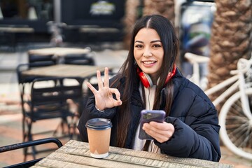 Young hispanic woman using smartphone sitting on the table doing ok sign with fingers, smiling friendly gesturing excellent symbol