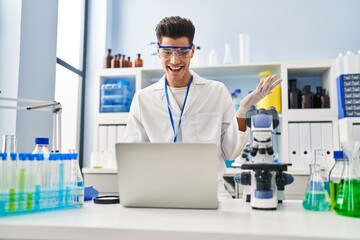 Young hispanic man working at scientist laboratory doing video call celebrating achievement with happy smile and winner expression with raised hand