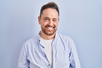 Middle age caucasian man standing over blue background winking looking at the camera with sexy expression, cheerful and happy face.