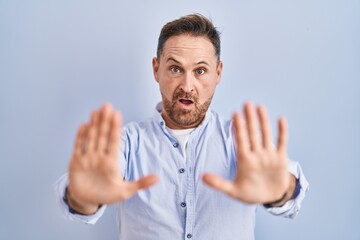 Fototapeta premium Middle age caucasian man standing over blue background doing stop gesture with hands palms, angry and frustration expression